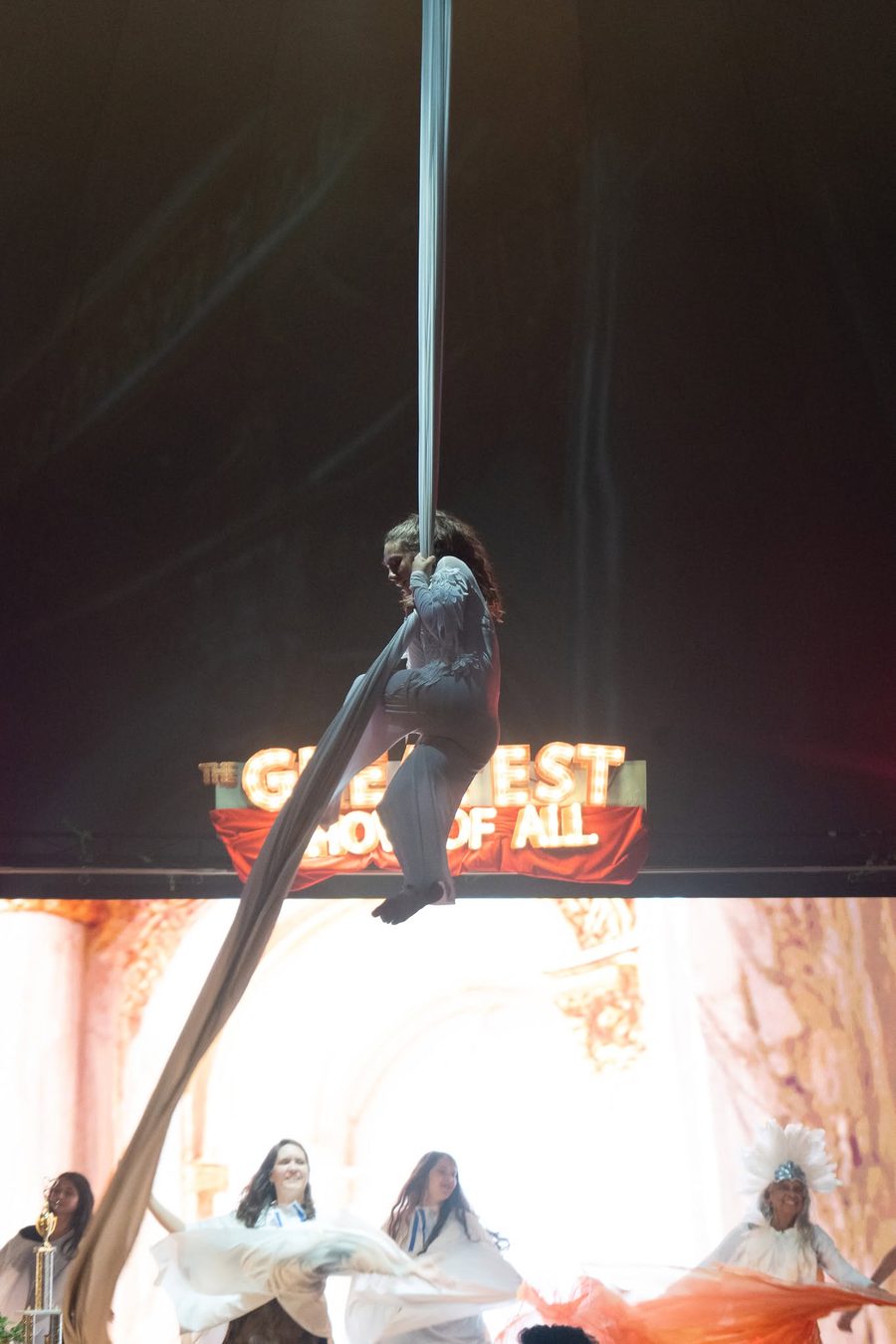 An aerialist performing on silks above the Greatest Show of All marquee