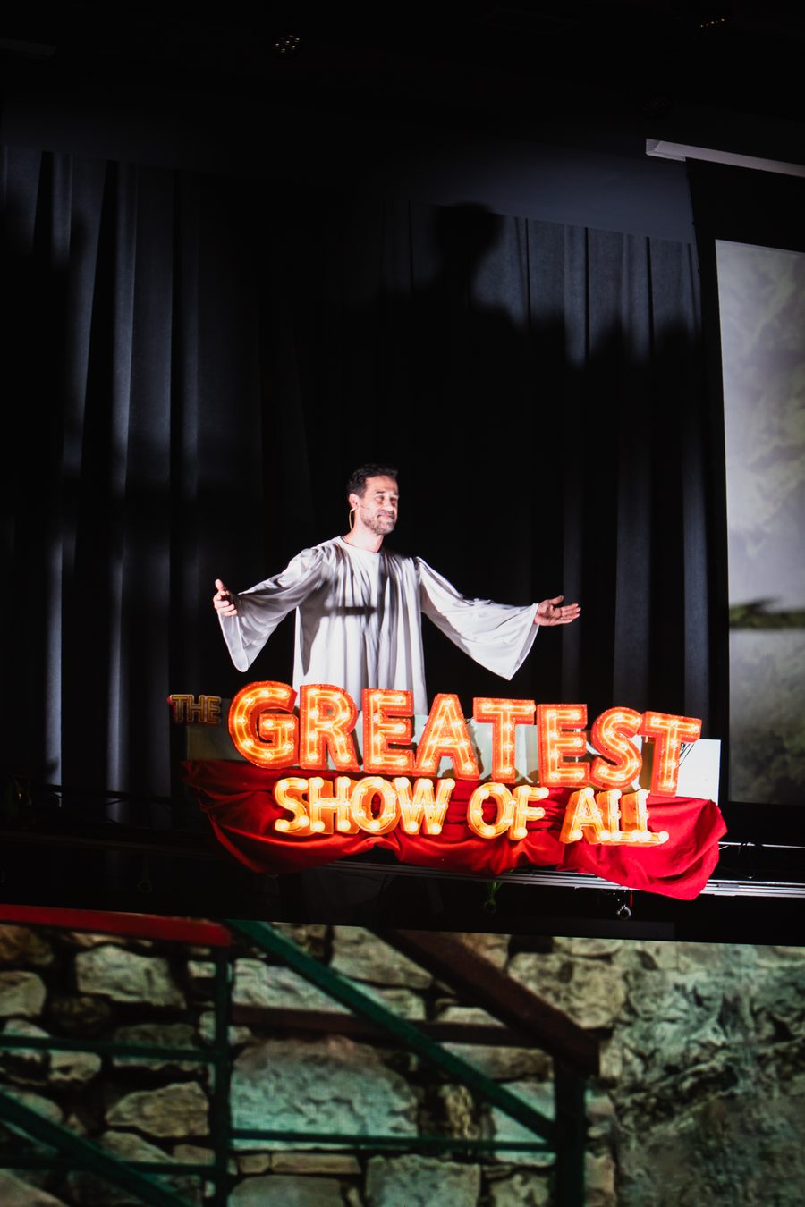 An actor with arms outstretched in front of the glowing Greatest Show of All sign